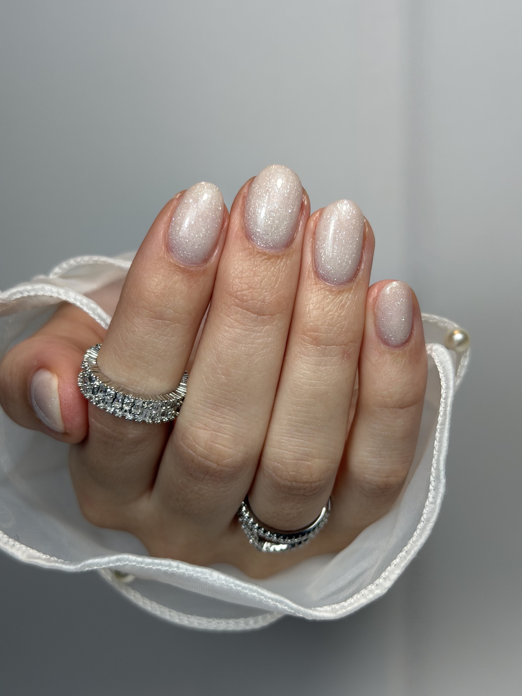 Beige nagels met glitter in amandelvorm, een hand met twee zilveren ringen in een wit doekje