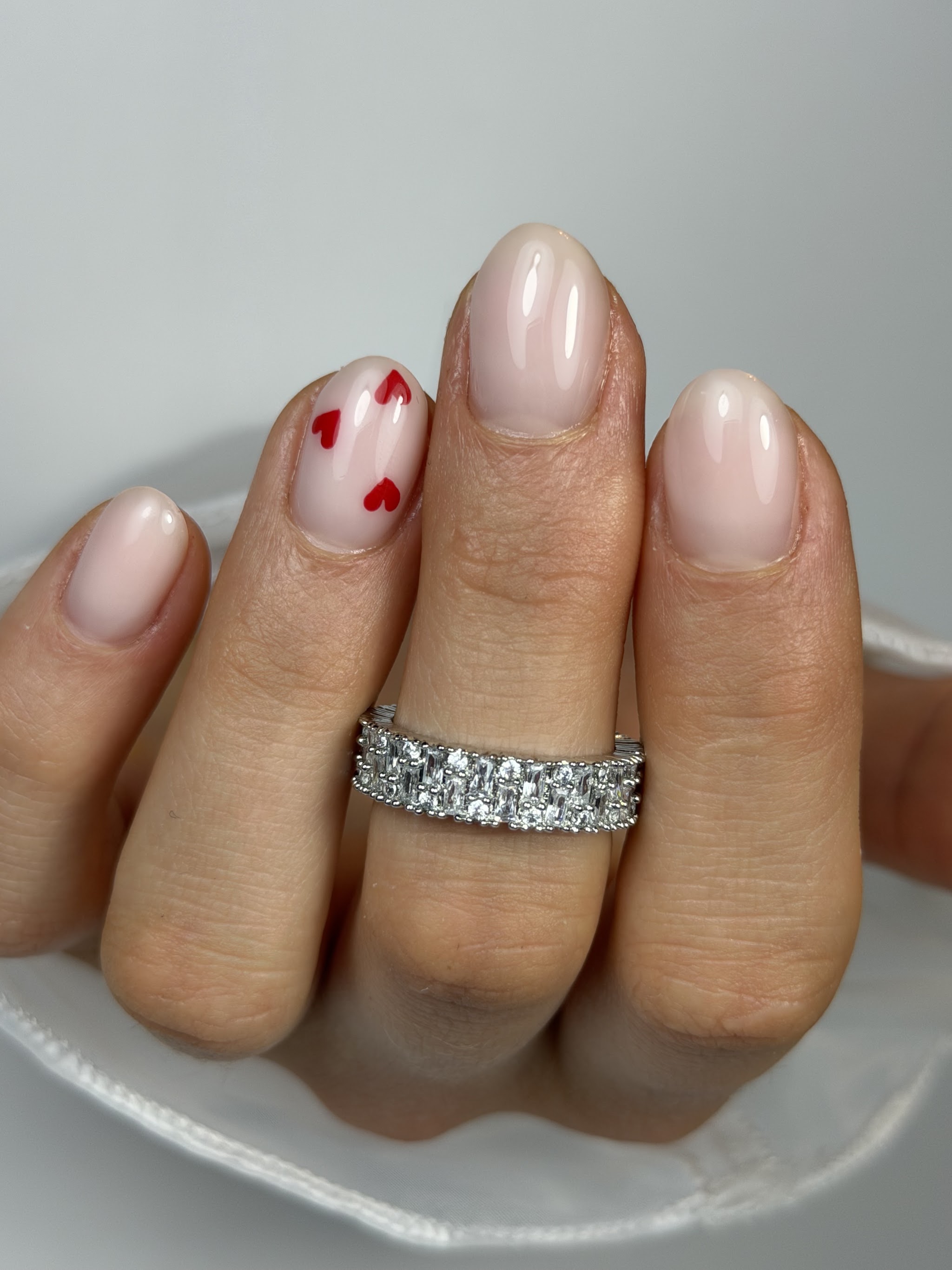 Nude nagels in amandelvorm met rode hartjes op de ringvinger, en een brede ring met steentjes